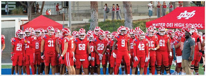 A group of people wearing red football uniforms and helmets stand together on a field near a red tent with “Mater Dei High School" written on it. Description generated by AI