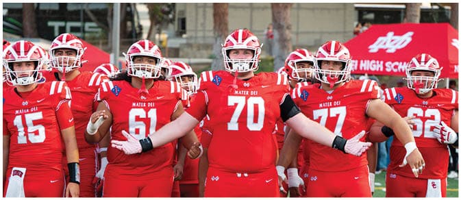 Two opposing American football teams in red and white uniforms are lined up facing each other on a field, preparing for a play. Description generated by AI