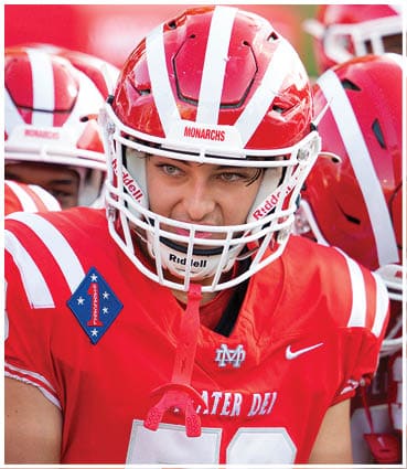 A person wearing a red and white football uniform and helmet with a focused expression is in the foreground while another person in similar gear is partially visible in the background. Description generated by AI