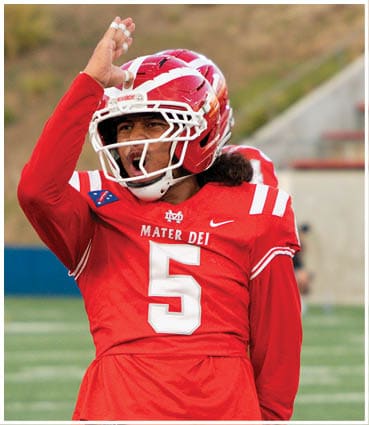 A person wearing a red football uniform with the number 5 and a helmet is celebrating with their arm raised on a football field. Description generated by AI