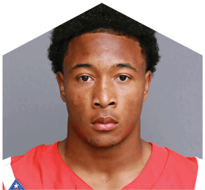 Person with short curly hair wearing a red sports jersey with a patch on the shoulder, looking directly at the camera against a plain gray background. Description generated by AI