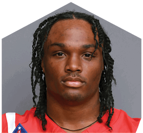 Person with braided hair wearing a red sports jersey looking directly at the camera against a plain gray background. Description generated by AI
