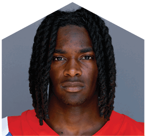 A person with shoulder-length dreadlocks wearing a red sports jersey looks directly at the camera against a plain gray background. Description generated by AI