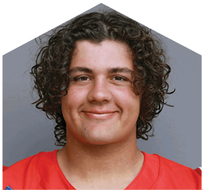 A person with curly hair is smiling and wearing a red sports jersey against a gray background. Description generated by AI