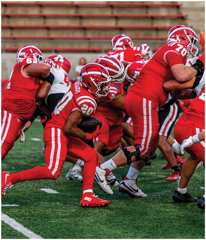 A group of football players in red uniforms are actively engaged in a play on a green field with empty bleachers in the background. Description generated by AI