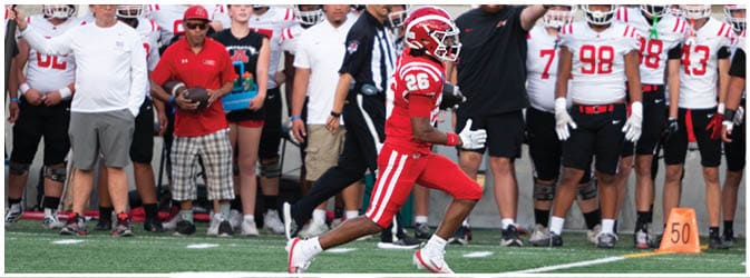 A group of people in football uniforms and coaches stand on the sidelines with one player in a red uniform running in front of them. Description generated by AI