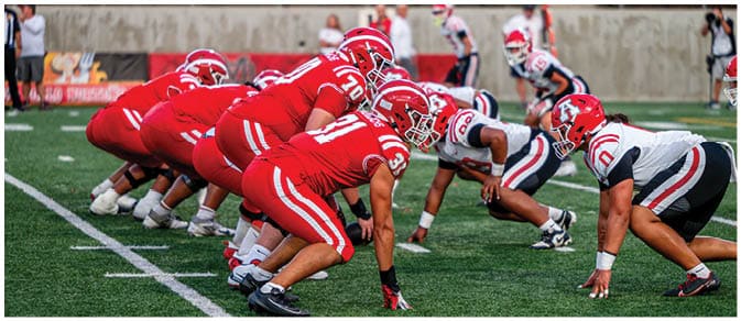 Two opposing American football teams are lined up facing each other on a green field, preparing for the next play. Description generated by AI