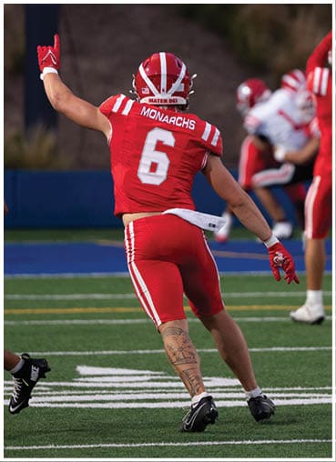 Two people in football uniforms are on a field with one raising an arm while another stands nearby, and a third person in the background also raises both arms. Description generated by AI