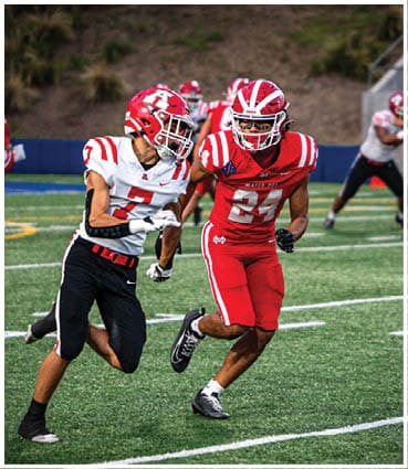 Two people in football uniforms are actively running on a field during a game, with one wearing red and the other wearing white and black. Description generated by AI