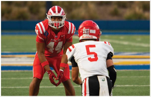 Two people in football uniforms and helmets face each other on a field, one in red and the other in white. Description generated by AI