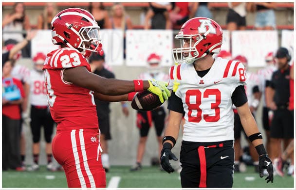 Two people in football uniforms are on a field, one wearing red and the other wearing white and black, with the person in red holding a football. Description generated by AI