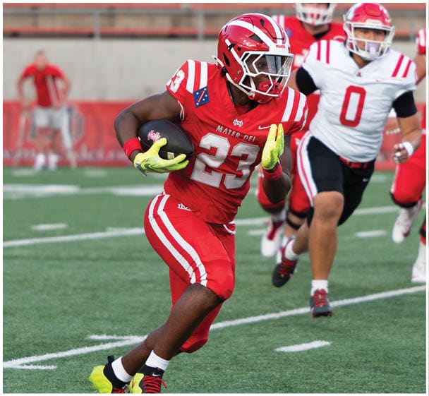An adult wearing a red football uniform with the number 23 is running with the ball while being chased by adults in white and red football uniforms on a field. Description generated by AI