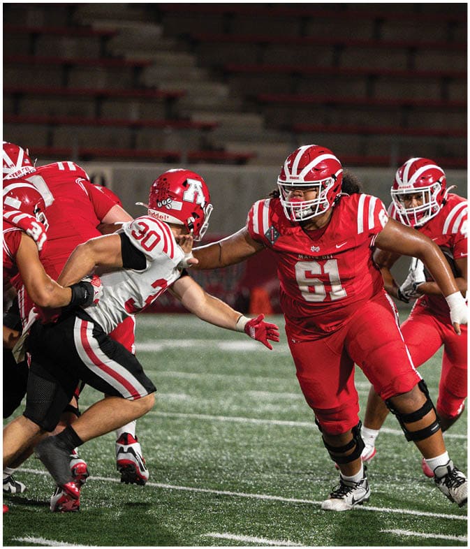 Four people wearing football uniforms are engaged in a play on a field with empty bleachers in the background. Description generated by AI