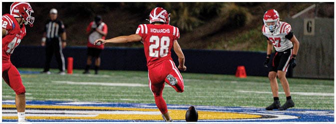 A person in a red football uniform is kicking a football on a field while two other people in sports attire watch. Description generated by AI