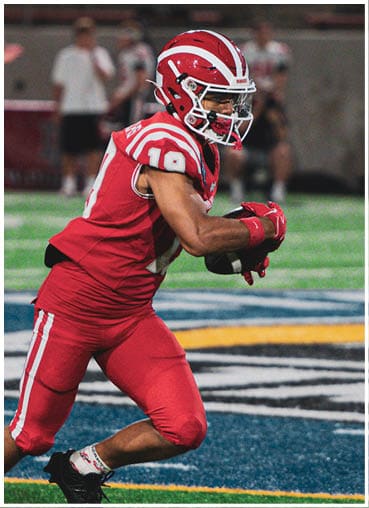A person wearing a red football uniform and helmet is running on a field during a game. Description generated by AI