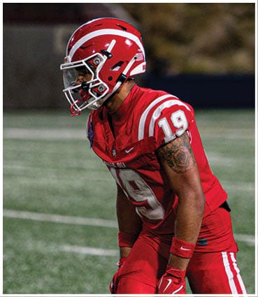 A person wearing a red football uniform with the number 19 and a matching helmet is standing on a football field. Description generated by AI