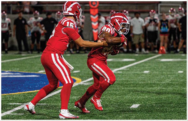 Two people in red football uniforms are on a field with one handing off the ball to the other during a game. Description generated by AI