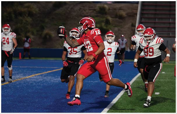 A person in red sports gear is running on a blue football field while holding a football, with several other people in white and black sports uniforms nearby. Description generated by AI