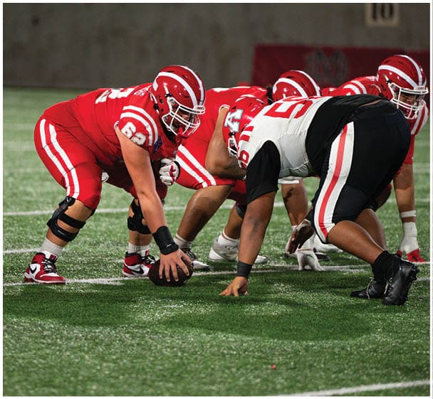 People in red and white football uniforms are lined up on a field preparing for a play against opponents in black and white uniforms. Description generated by AI