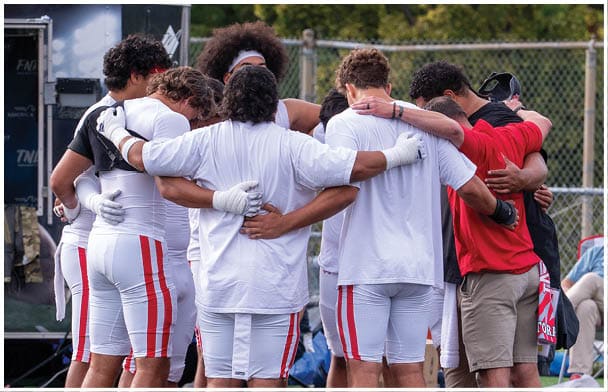 A group of people wearing sports attire and gloves stand in a close huddle on a field with their arms around each other. Description generated by AI