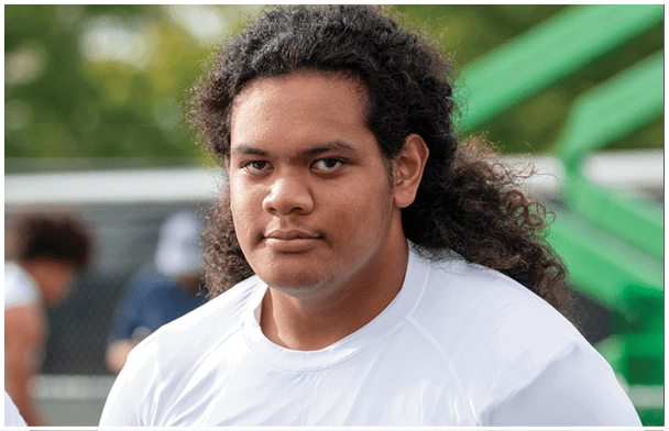 A person with long curly hair wearing a white shirt is looking directly at the camera with a neutral expression while two other people are blurred in the background near a chain-link fence. Description generated by AI