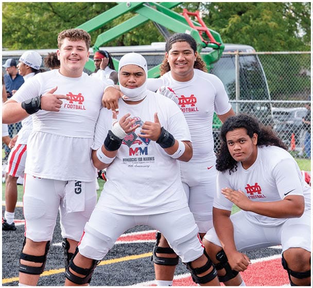 Four people wearing white football gear pose together on a sports field with a chain-link fence and equipment in the background. Description generated by AI