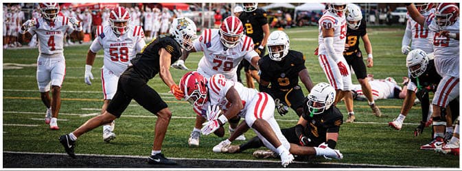 A group of people in football uniforms are actively engaged in a game on a field with a scoreboard visible in the background. Description generated by AI