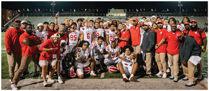 A large group of people wearing football uniforms and red team apparel pose together on a football field at night with stadium lights and empty bleachers in the background. Description generated by AI