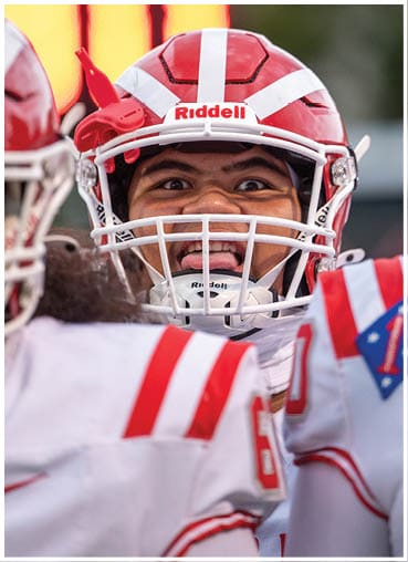 A person wearing a red and white football helmet and uniform is making a playful face with their tongue out while interacting with teammates. Description generated by AI