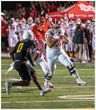 A person in white football gear with a knee brace is running with the ball while a person in black football gear attempts to tackle them on a field with spectators and team members in the background. Description generated by AI