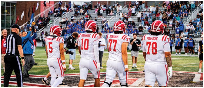 Four people in white and red football uniforms stand on a field facing away from the camera with a crowd seated in bleachers in the background. Description generated by AI