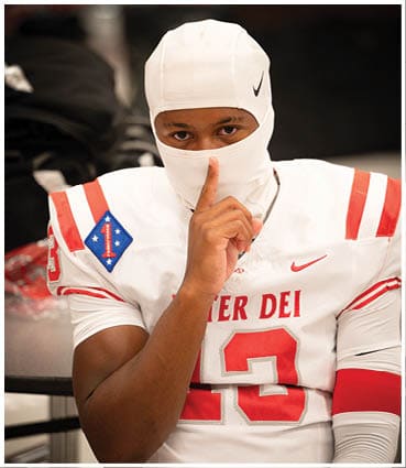 A person wearing a white football uniform and helmet liner with a finger held up to their lips in a shushing gesture. Description generated by AI