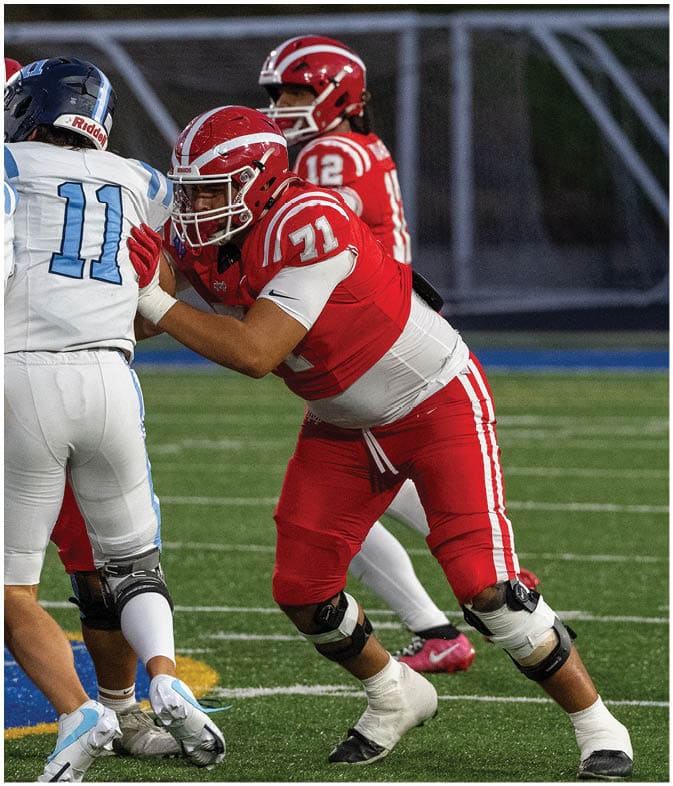 Two people in red football uniforms and helmets are positioned on a field, appearing ready for a play. Description generated by AI