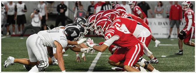 Two opposing American football teams in red and white uniforms are lined up facing each other on a field, ready for the next play. Description generated by AI