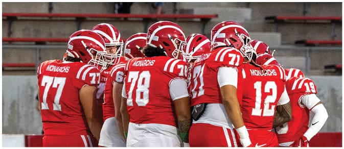 A group of people wearing red football uniforms and helmets are gathered closely together on a field with empty bleachers in the background. Description generated by AI