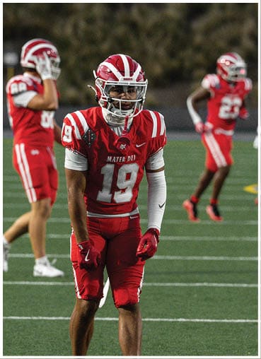 A person in a red and white football uniform with the number 19 stands on a field with two other players in similar uniforms in the background. Description generated by AI