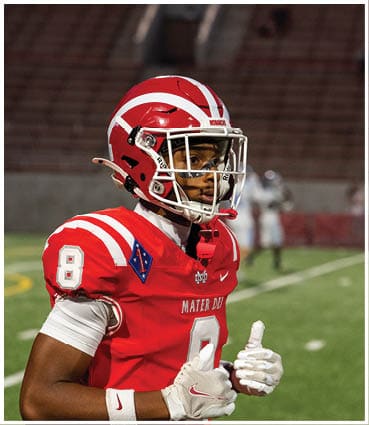 A person wearing a red and white football helmet and red jersey with the number 8 stands on a football field with blurred stadium seats in the background. Description generated by AI