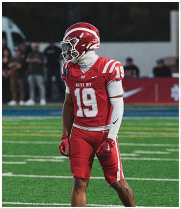 A person wearing a red football uniform with the number 19 stands on a green field with white yard lines and a blurred crowd in the background. Description generated by AI