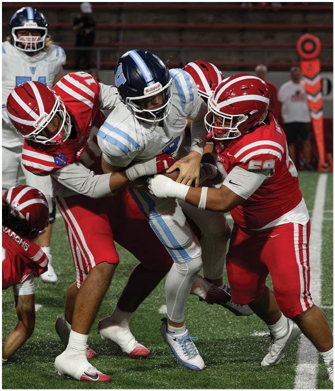People in football uniforms are engaged in a dynamic play on a field with one person in red falling while others in white and blue attempt to block or tackle. Description generated by AI