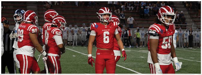 People wearing red and white football uniforms stand on a grass field during a game or practice. Description generated by AI