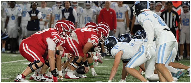 Two opposing American football teams in red and white uniforms are lined up facing each other on the field, preparing for a play. Description generated by AI