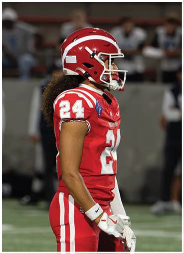 A person wearing a red football uniform with the number 24 and a matching red helmet stands on a field with blurred spectators in the background. Description generated by AI