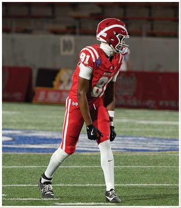A person wearing a red and white football uniform with the number 81 is standing on a football field in a ready position. Description generated by AI