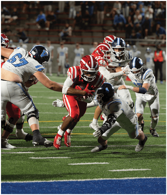 A group of people wearing football uniforms are actively playing on a field with one person in red running with the ball while others in white and blue attempt to tackle or block. Description generated by AI