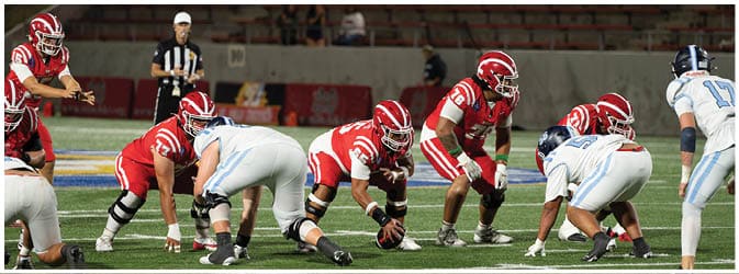 People in football uniforms are lined up on a field preparing for a play during a game. Description generated by AI