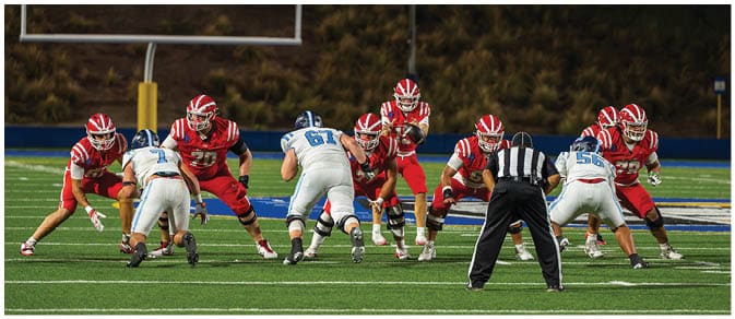 Two opposing American football teams in red and white uniforms are lined up facing each other on a field with a referee standing behind the team in white. Description generated by AI