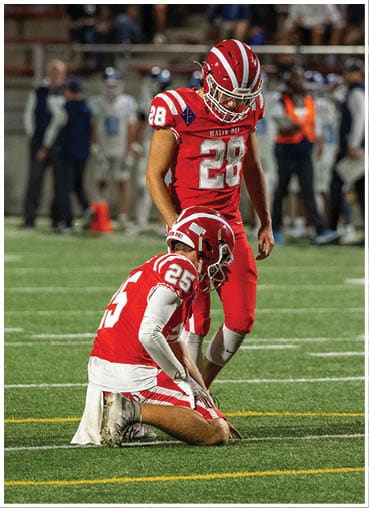 Three football players in red uniforms are on a field with one player kneeling and holding the ball while the other two stand nearby. Description generated by AI