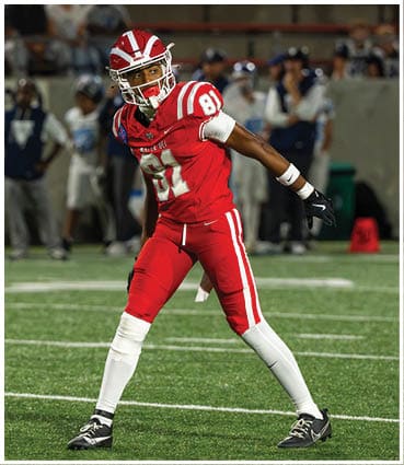 A person in a red football uniform is preparing to throw a football on a green field with other players and spectators in the background. Description generated by AI