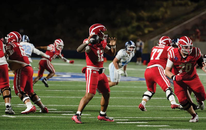 A group of people in red and white football uniforms are playing on a field at night with one person preparing to throw a football. Description generated by AI