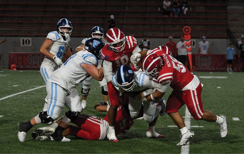 Players in red and white uniforms tackle a player in a white and blue uniform during a football game on a green field. Description generated by AI